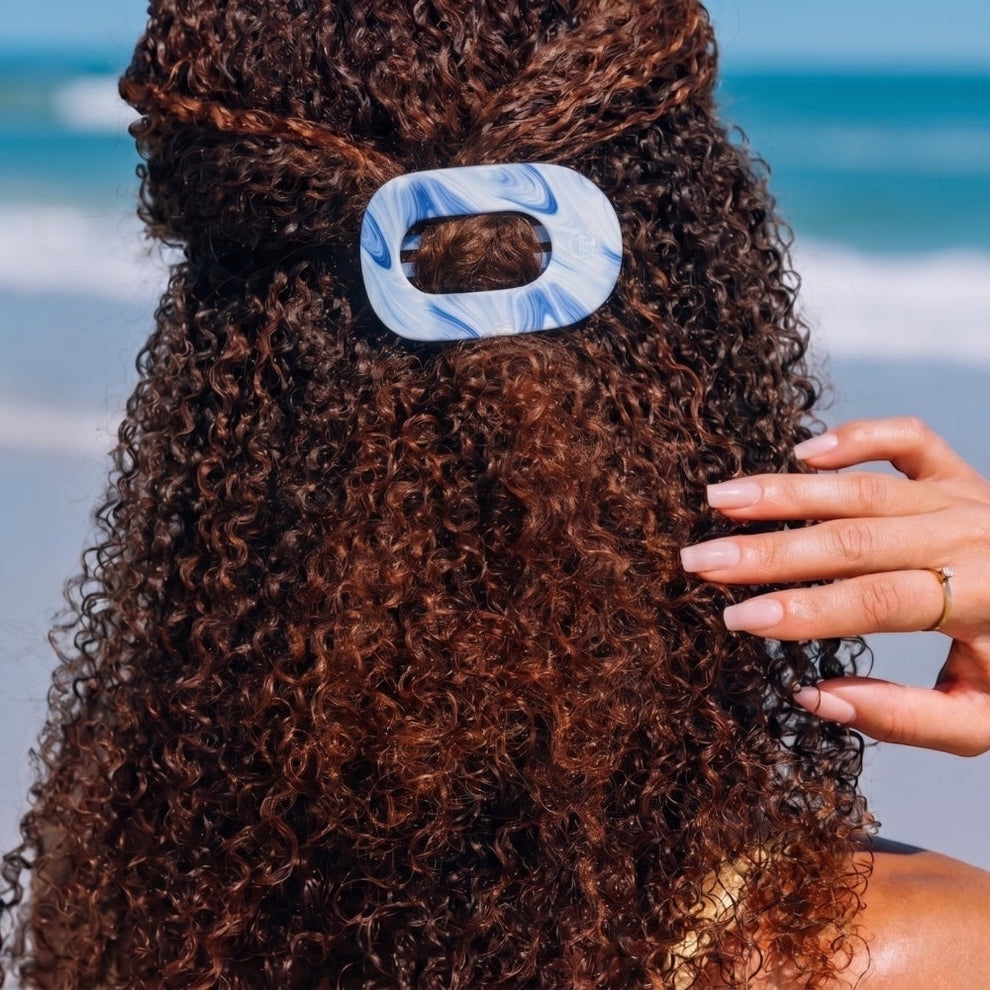 TELETIES blue and white marbled flat round hair clip on curly brown hair at the beach.
