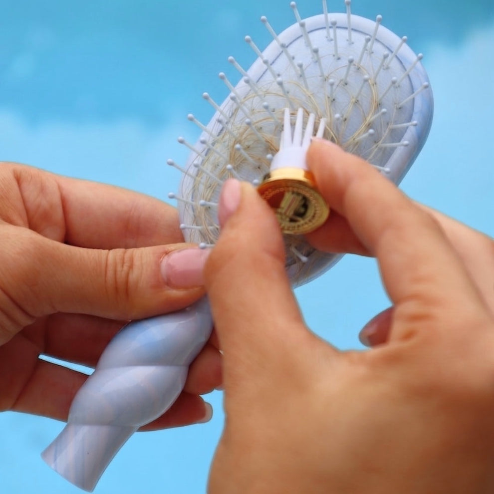 TELETIES light blue detangler hair brush with a unique swirl pattern handle, being cleaned with a small tool.
