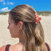 TELETIES Just Keep Swimming Medium Claw Clip in pink and orange marbled resin, holding back blonde hair on a beach.
