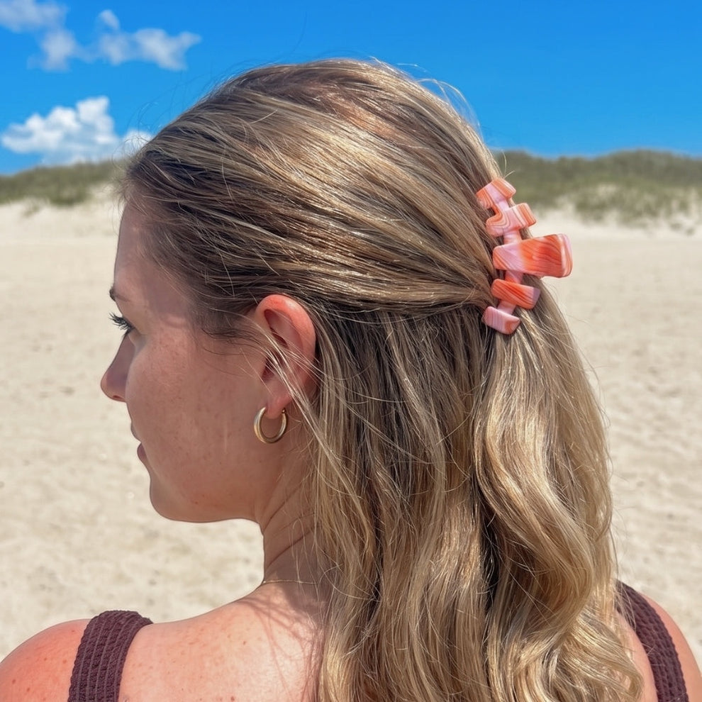 TELETIES Just Keep Swimming Medium Claw Clip in pink and orange marbled resin, holding back blonde hair on a beach.
