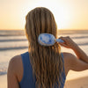 TELETIES mini detangler hair brush with blue tie-dye pattern and gold trim, used on wet hair at the beach.
