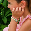 A young girl wearing TELETIES Pua and Heihei hair ties as bracelets on her wrist against a green leafy background.
