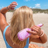 TELETIES pink and white tie-dye detangler hair brush with gold trim, used by a blonde woman on the beach.
